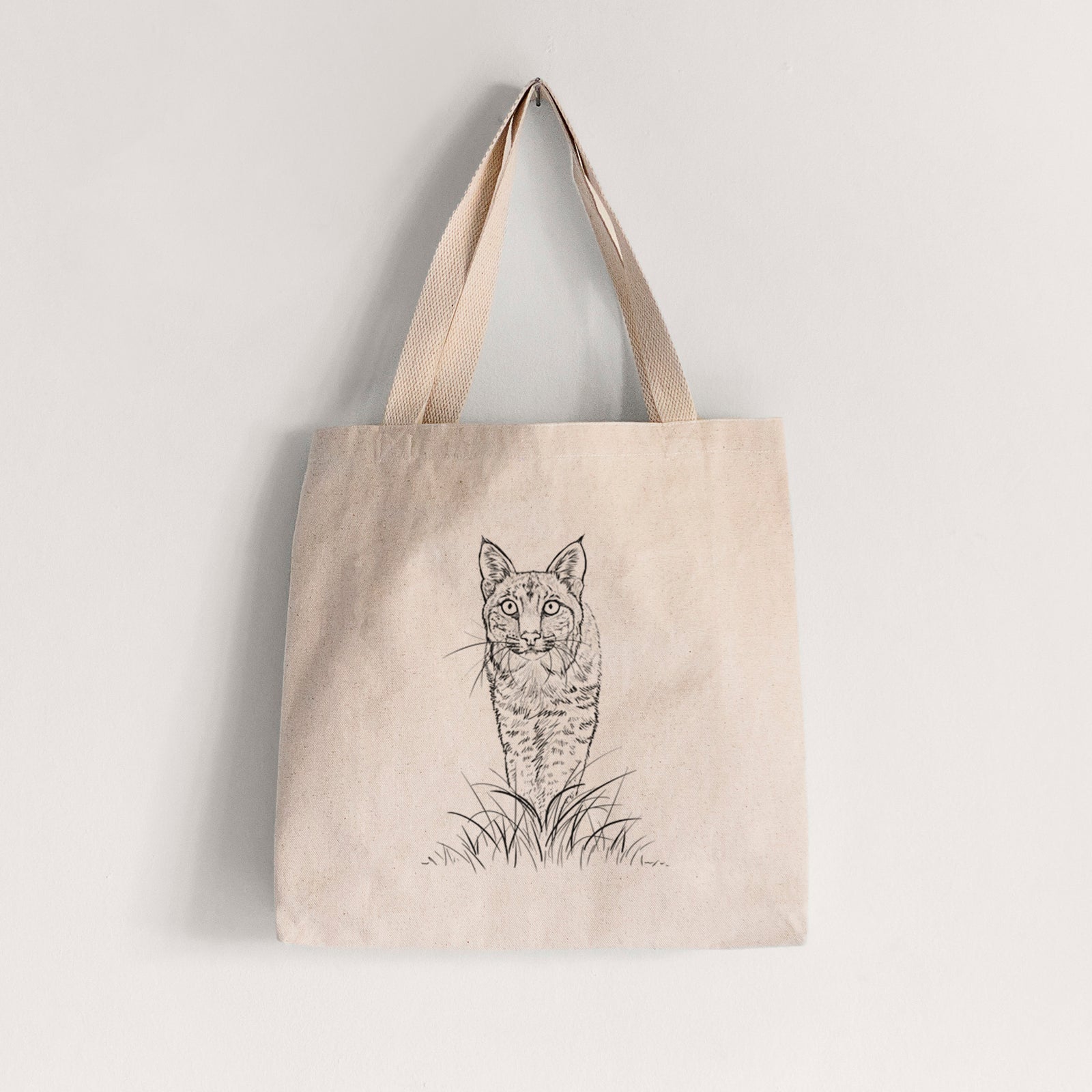 Natural canvas tote bag printed with a black ink illustration of a bobcat standing in tall grass, facing forward.