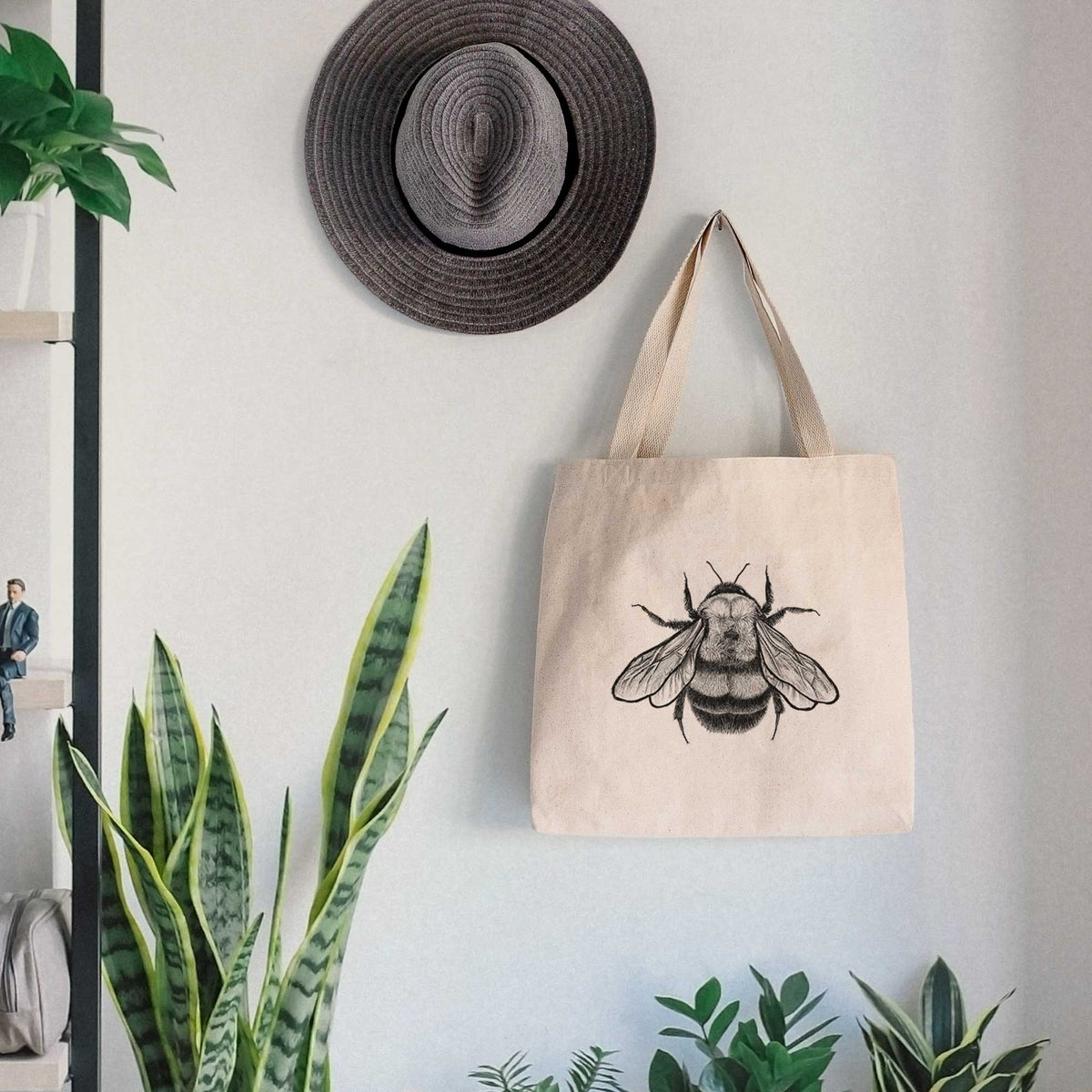 Canvas tote bag printed with a detailed black ink illustration of a rusty patched bumble bee with its wings spread, seen from above, hanging on a wall hook below a hat, next to potted plants.