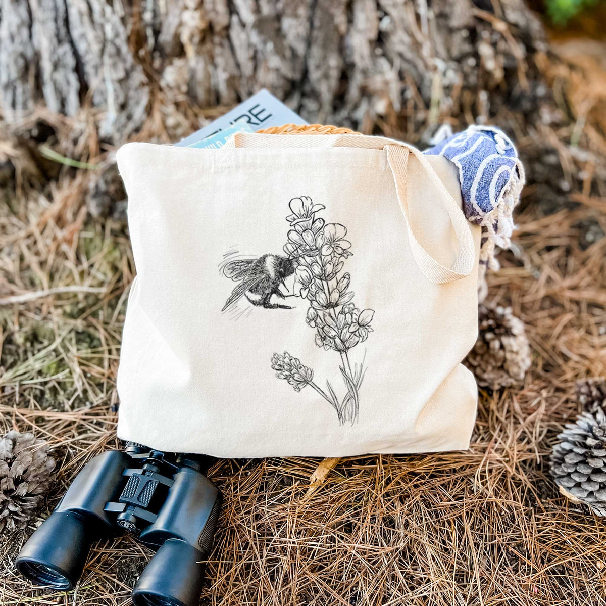 Canvas tote bag printed with a black ink illustration of a bumble bee visiting a sprig of lavender, resting on dry grass beside a pair of binoculars.
