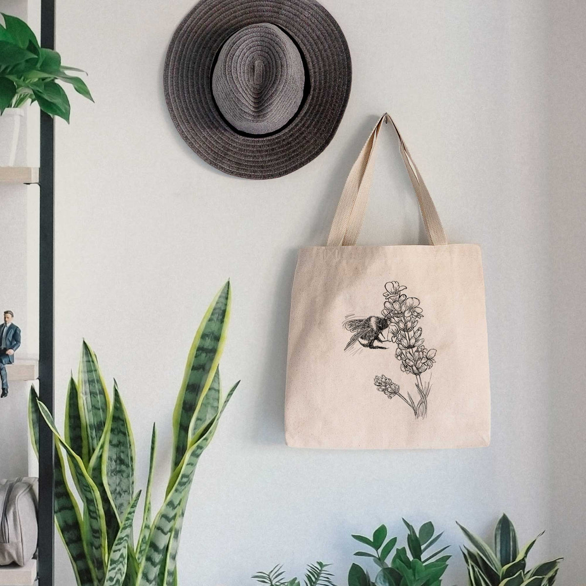 Canvas tote bag printed with a black ink illustration of a bumble bee visiting a sprig of lavender, hanging on a wall hook below a hat, next to potted plants.