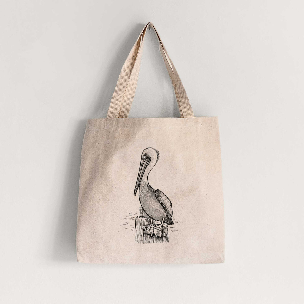 Natural canvas tote bag printed with a detailed black ink illustration of a brown pelican perched on a wooden piling.