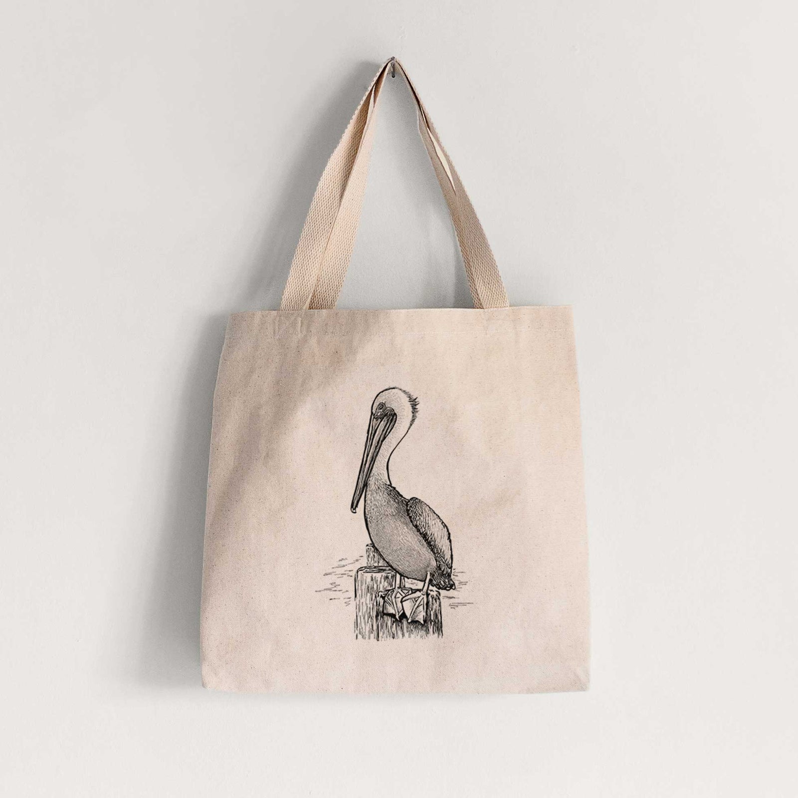 Natural canvas tote bag printed with a detailed black ink illustration of a brown pelican perched on a wooden piling.