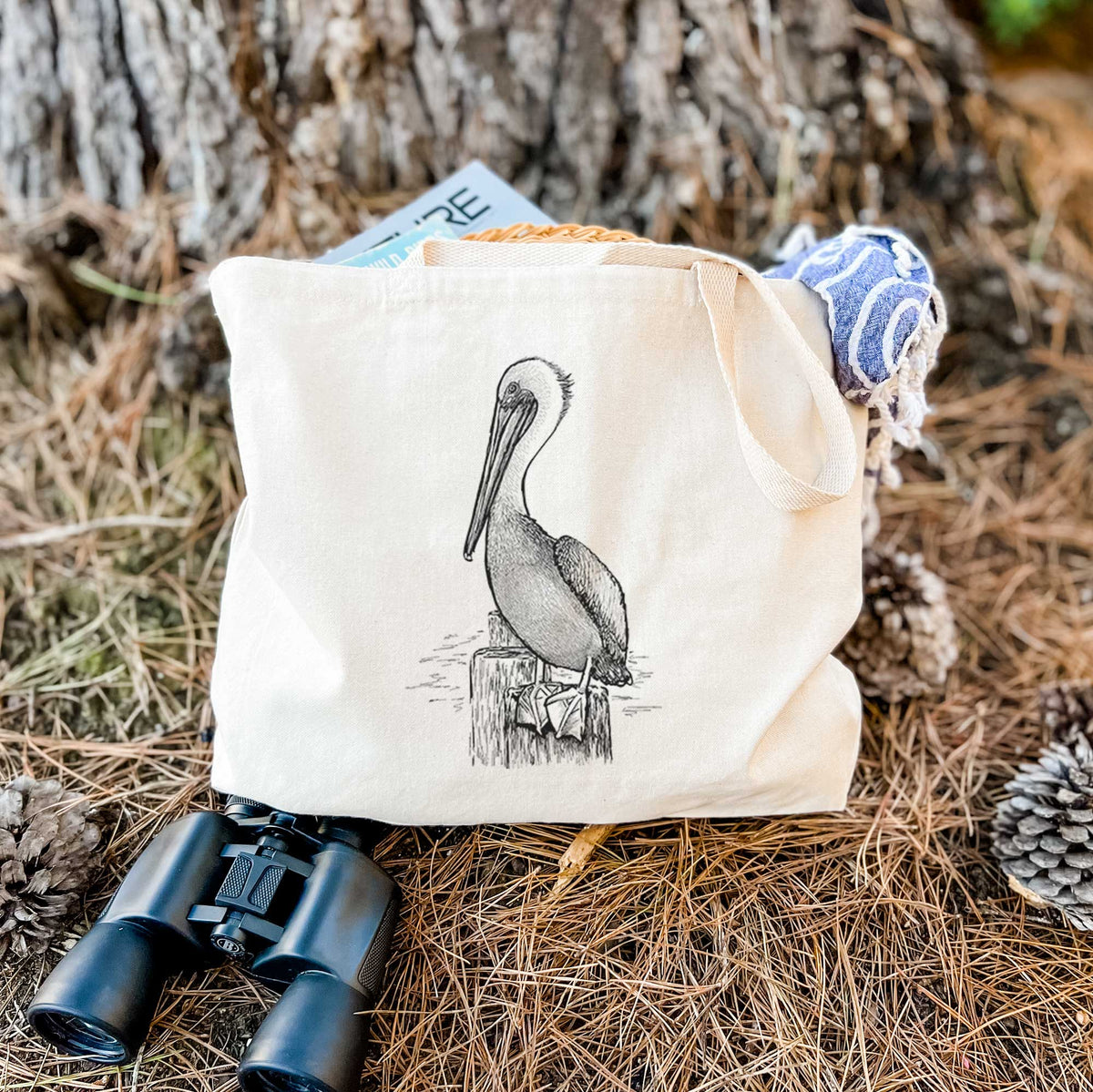 Canvas tote bag printed with a detailed black ink illustration of a brown pelican perched on a wooden piling, resting on dry grass beside a pair of binoculars.