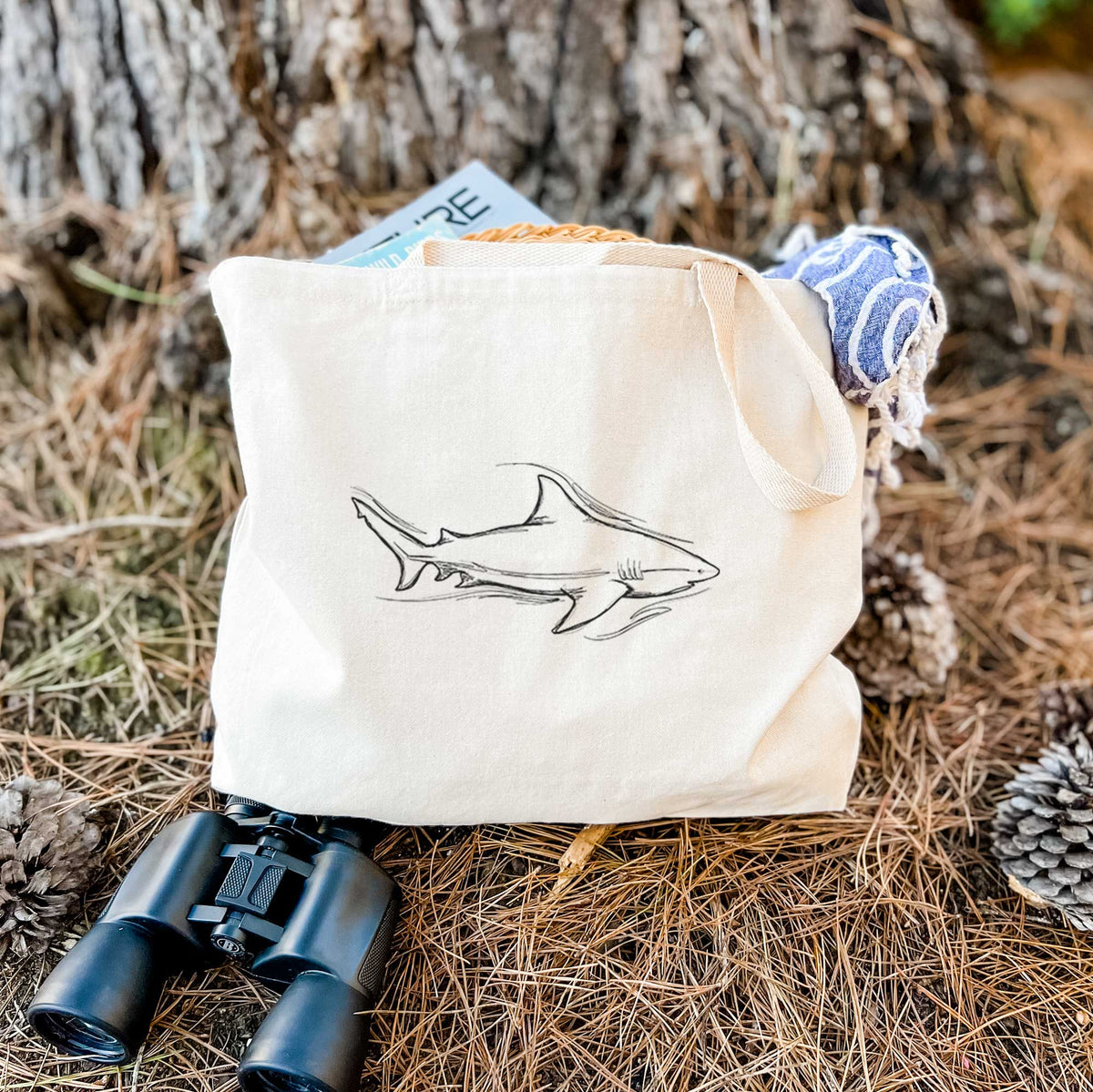 Canvas tote bag printed with a black ink illustration of a bull shark swimming, resting on dry grass beside a pair of binoculars.
