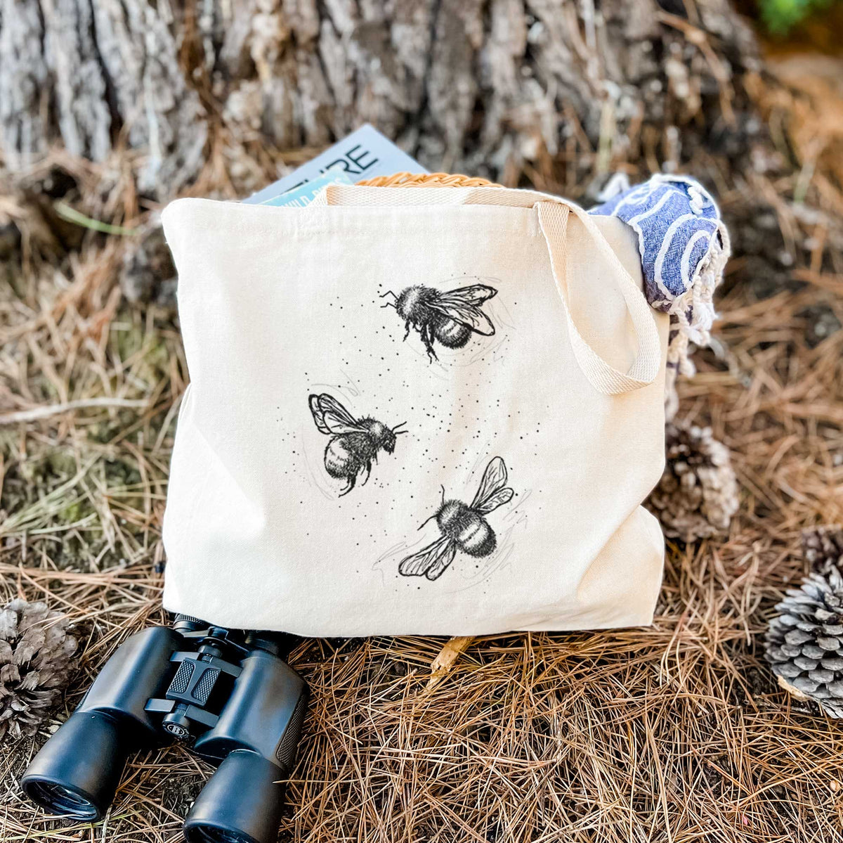 Canvas tote bag printed with a black ink illustration of three bumble bees flying, resting on dry grass beside a pair of binoculars.