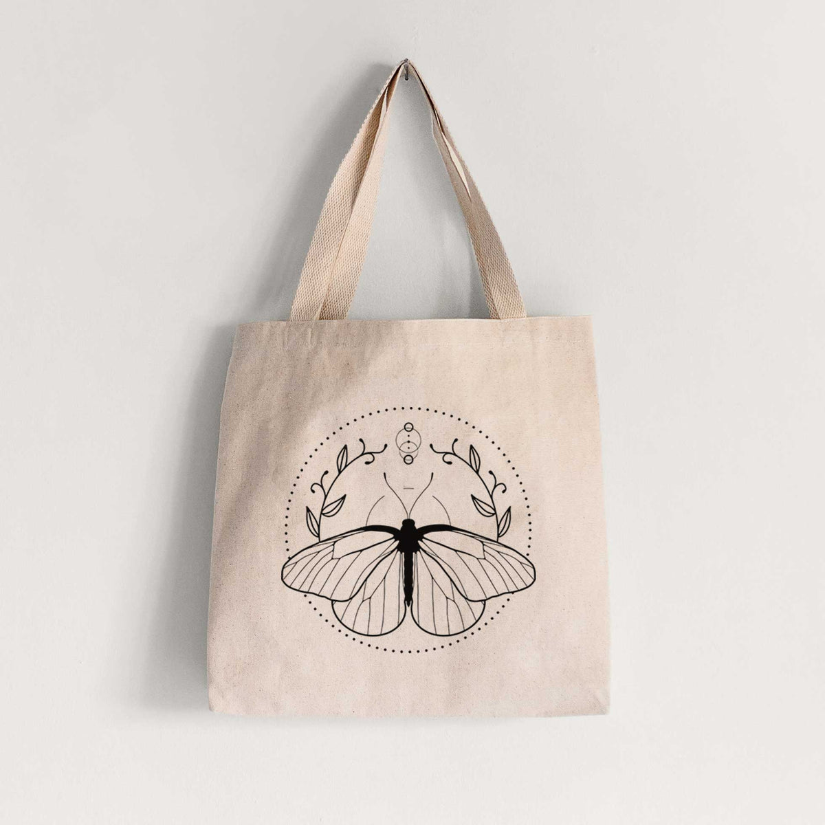Natural canvas tote bag printed with a black ink butterfly inside a circle, framed by moon phases and leaves.