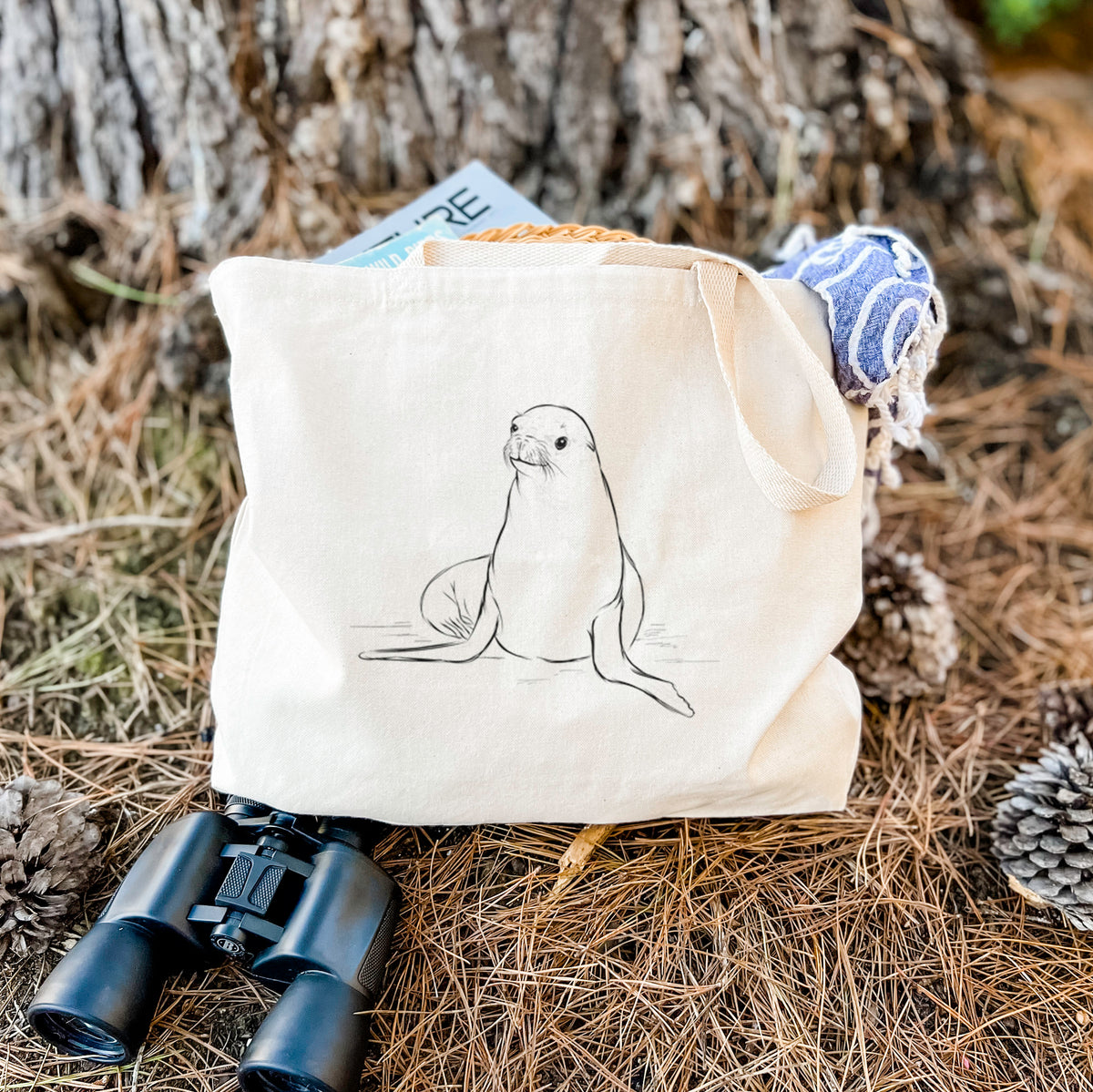 Canvas tote bag printed with a black ink illustration of a California sea lion sitting upright, resting on dry grass beside a pair of binoculars.