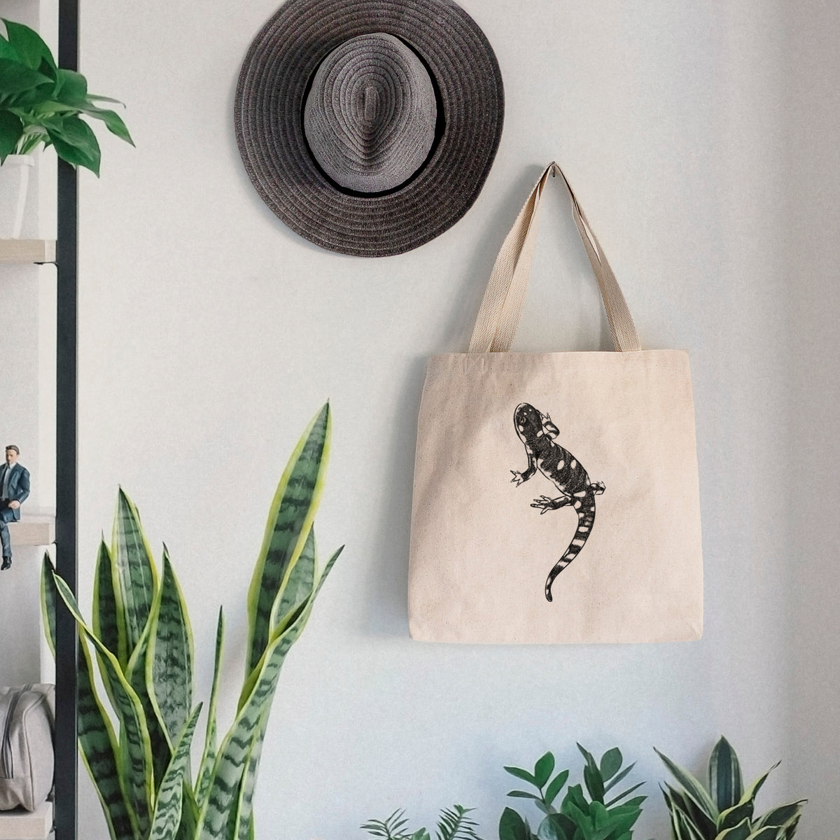 Canvas tote bag printed with a detailed black ink illustration of a spotted California tiger salamander, hanging on a wall hook below a hat, next to potted plants.