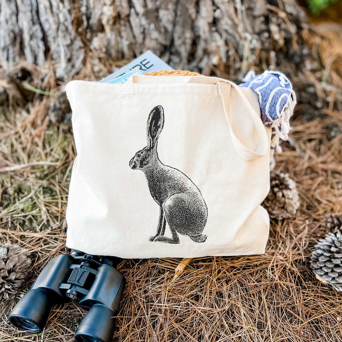 Canvas tote bag printed with a detailed black ink illustration of a black-tailed jackrabbit sitting in profile with tall ears, resting on dry grass beside a pair of binoculars.