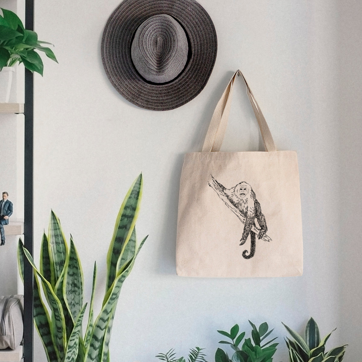 Canvas tote bag printed with a detailed black ink illustration of a capuchin monkey climbing a branch, hanging on a wall hook below a hat, next to potted plants.