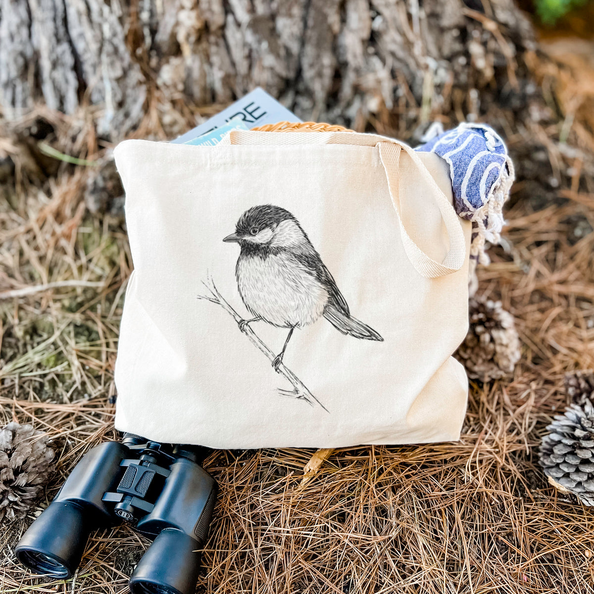 Canvas tote bag printed with a detailed black ink sketch of a black-capped chickadee perched on a branch, resting on dry grass beside a pair of binoculars.