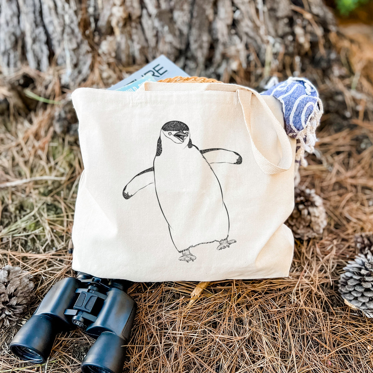 Canvas tote bag printed with a black ink illustration of a chinstrap penguin standing with its flippers out, resting on dry grass beside a pair of binoculars.