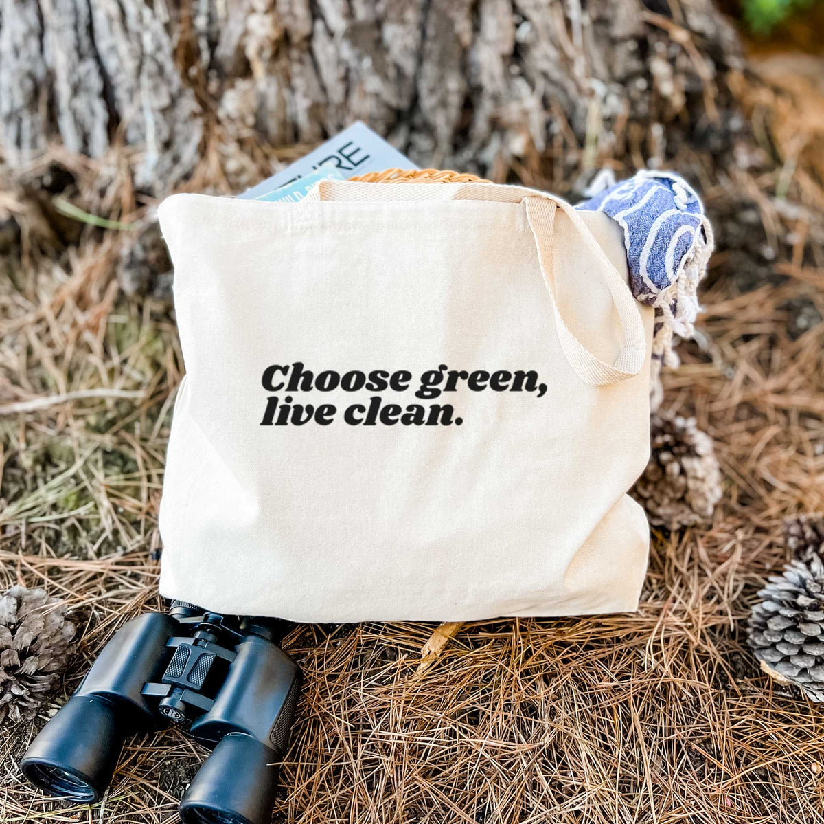 Canvas tote bag printed with small black script words reading Choose Green, Live Clean on the upper chest, resting on dry grass beside a pair of binoculars.