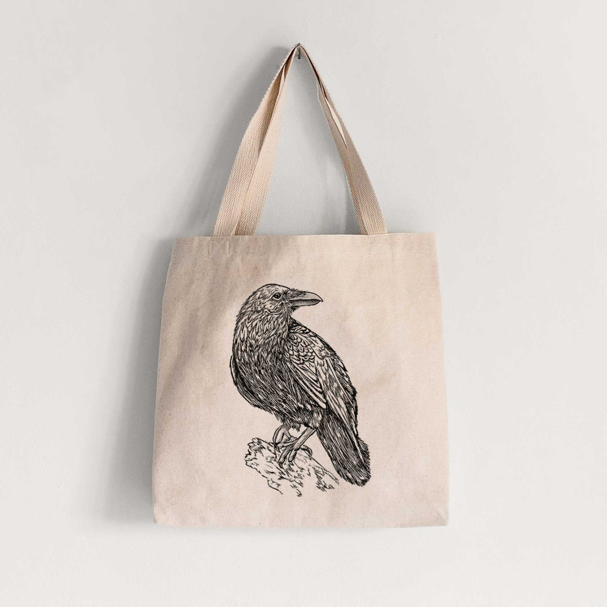 Natural canvas tote bag printed with a detailed black ink sketch of a common raven perched on a rock.