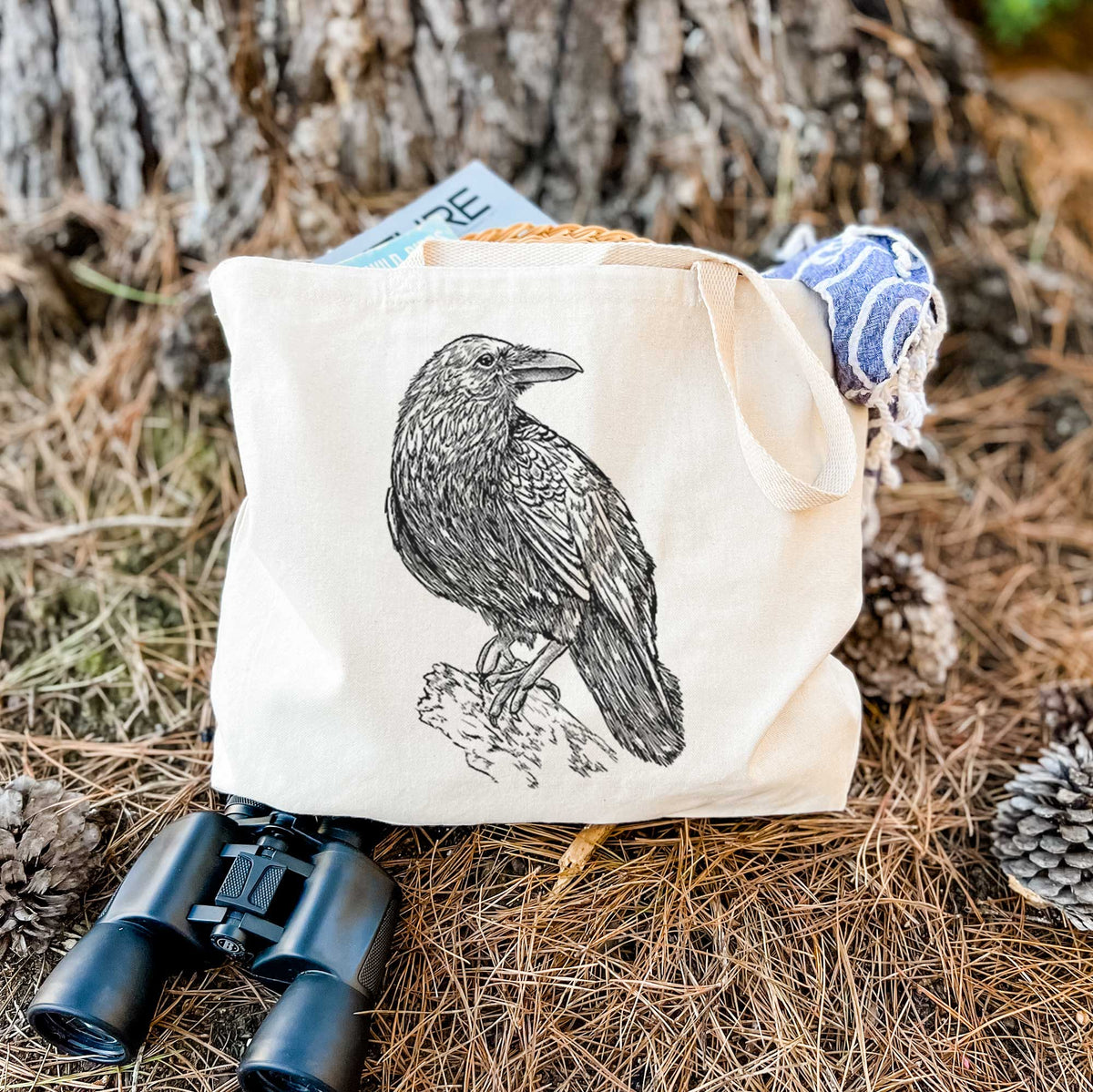 Canvas tote bag printed with a detailed black ink sketch of a common raven perched on a rock, resting on dry grass beside a pair of binoculars.