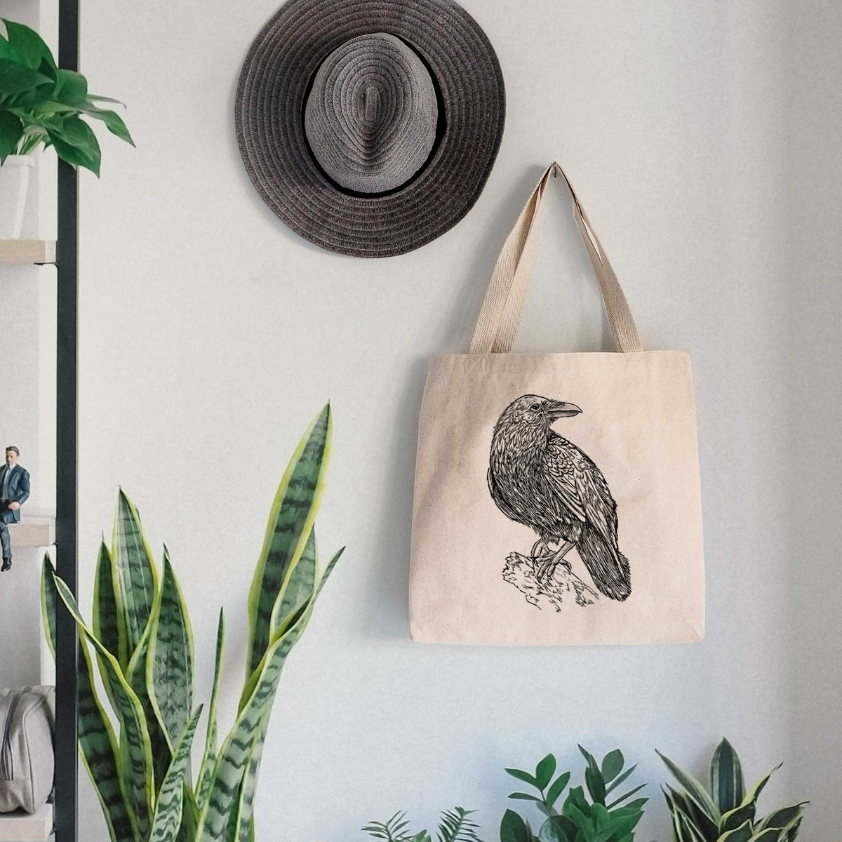 Canvas tote bag printed with a detailed black ink sketch of a common raven perched on a rock, hanging on a wall hook below a hat, next to potted plants.