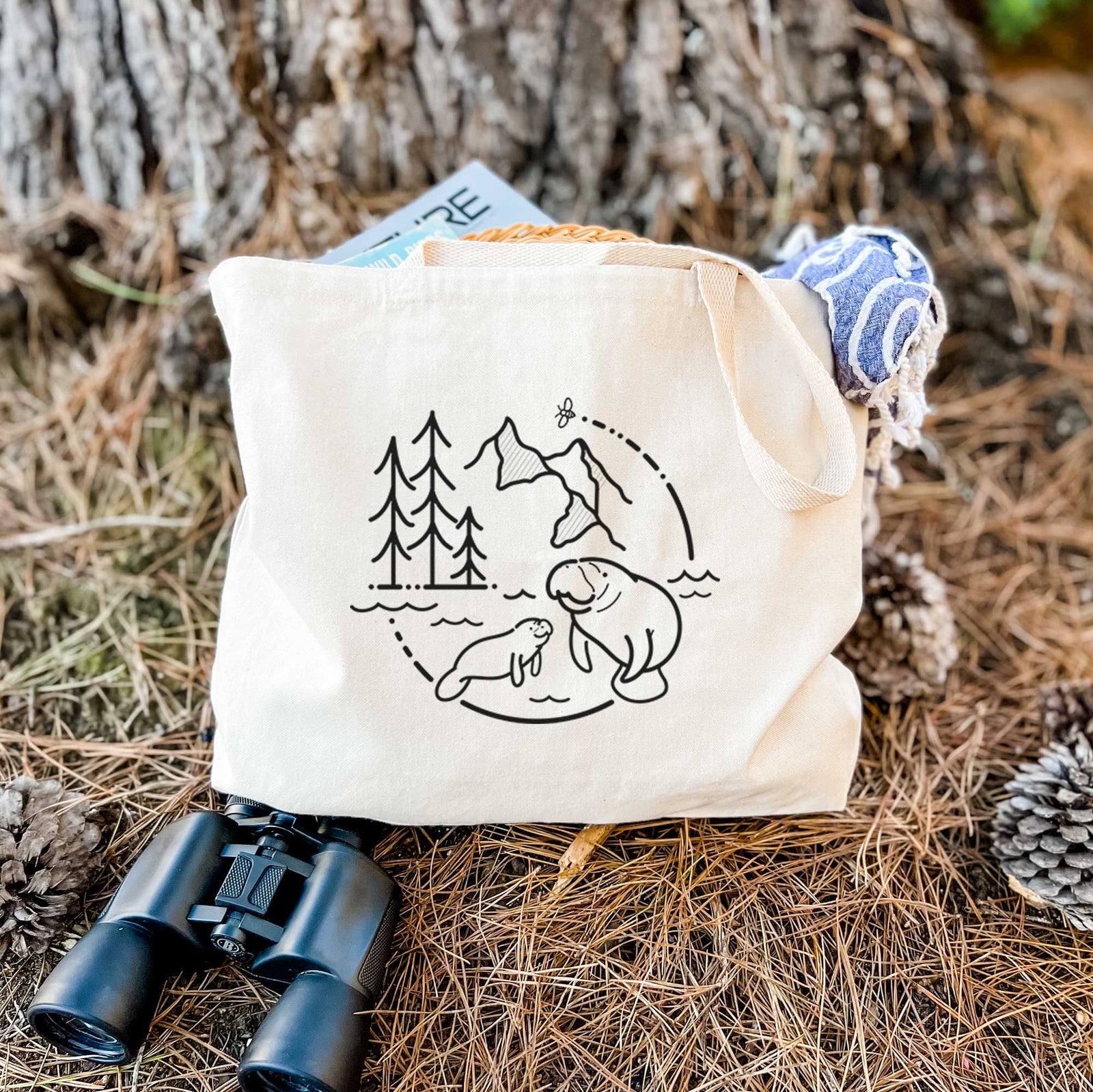 Natural canvas tote bag printed with a black line art circle connecting pine trees, mountains, a bee and two manatees swimming in water.