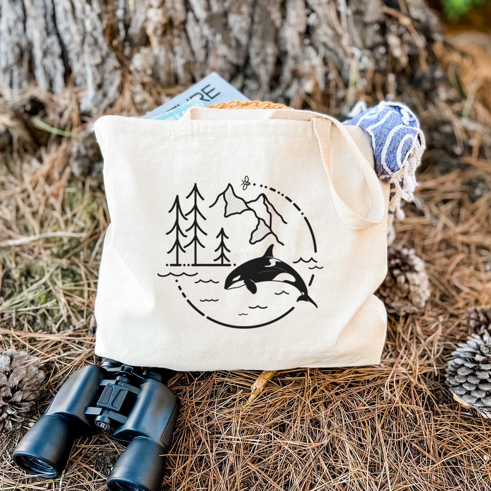 Natural canvas tote bag printed with a black line art circle connecting pine trees, mountains, a bee and an orca leaping from ocean waves.