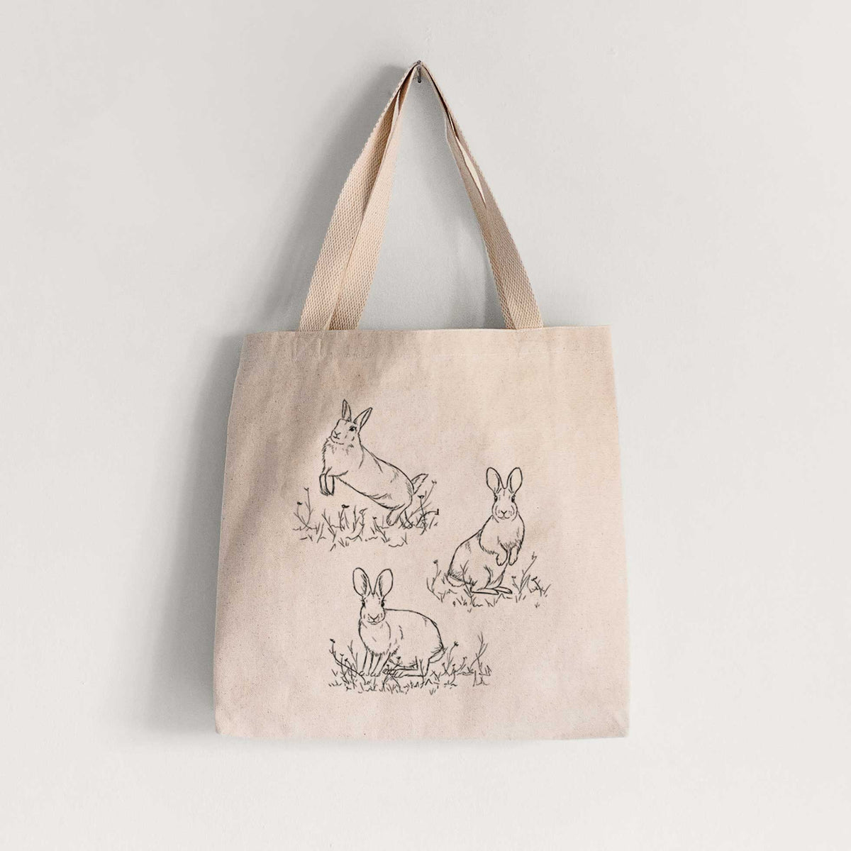 Natural canvas tote bag printed with a black ink illustration of three cottontail rabbits among grass and flowers.