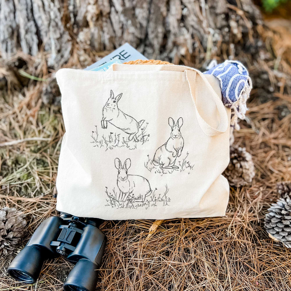 Canvas tote bag printed with a black ink illustration of three cottontail rabbits among grass and flowers, resting on dry grass beside a pair of binoculars.