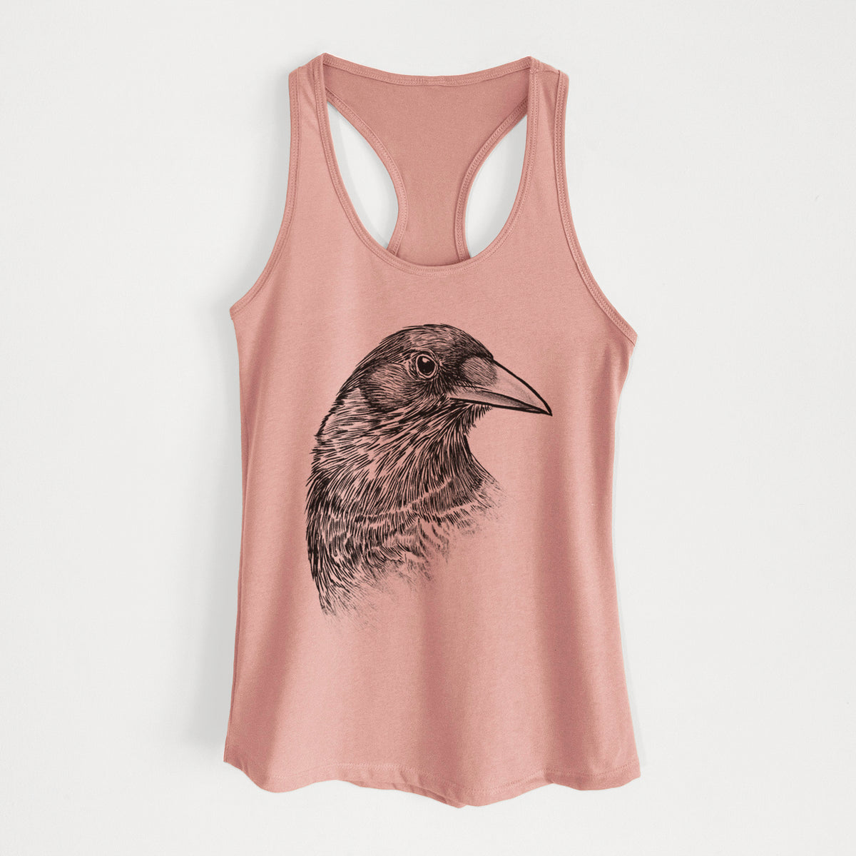 Desert pink women's racerback tank top laid flat, printed with a detailed black ink portrait of a crow's head in profile.