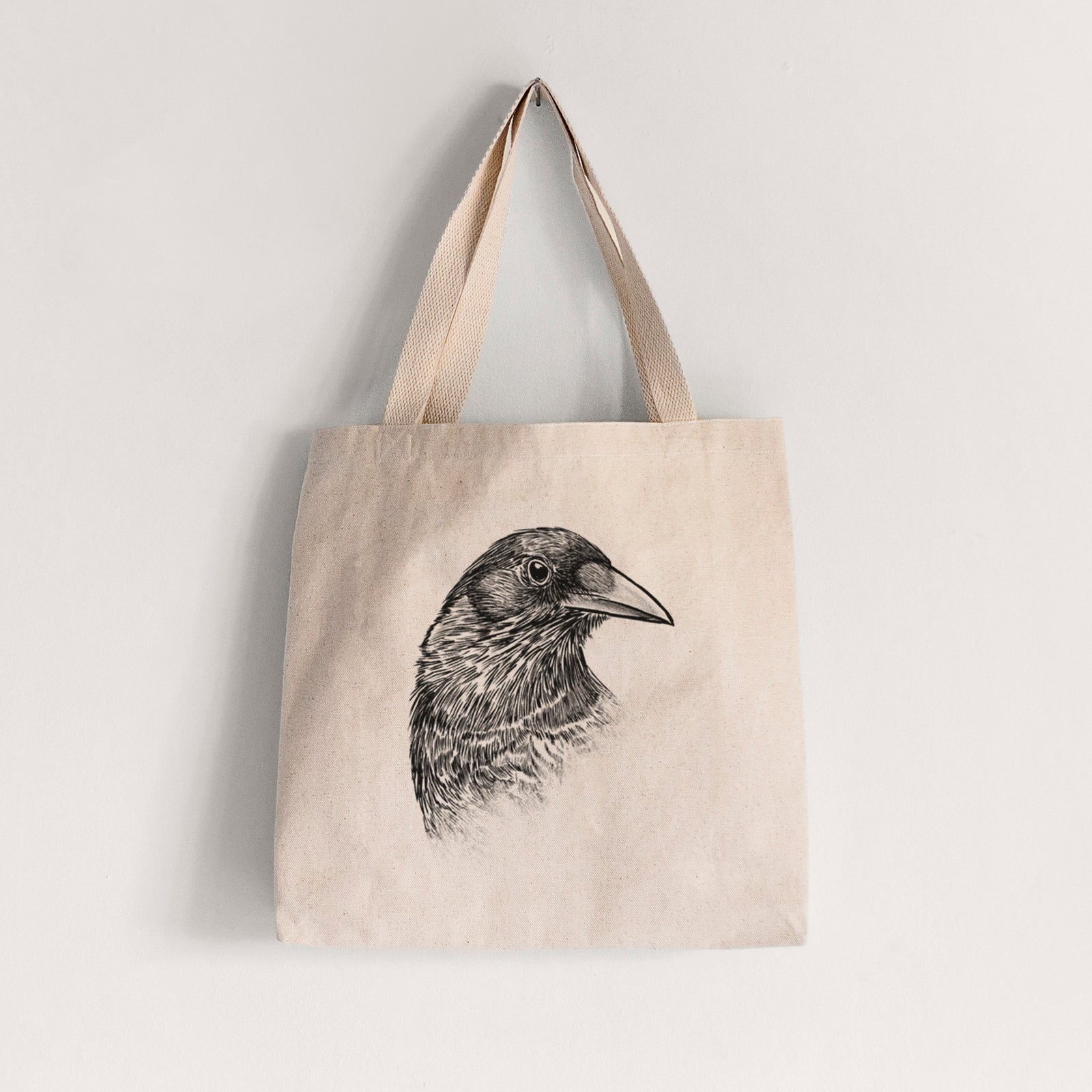 Natural canvas tote bag printed with a detailed black ink portrait of a crow's head in profile.