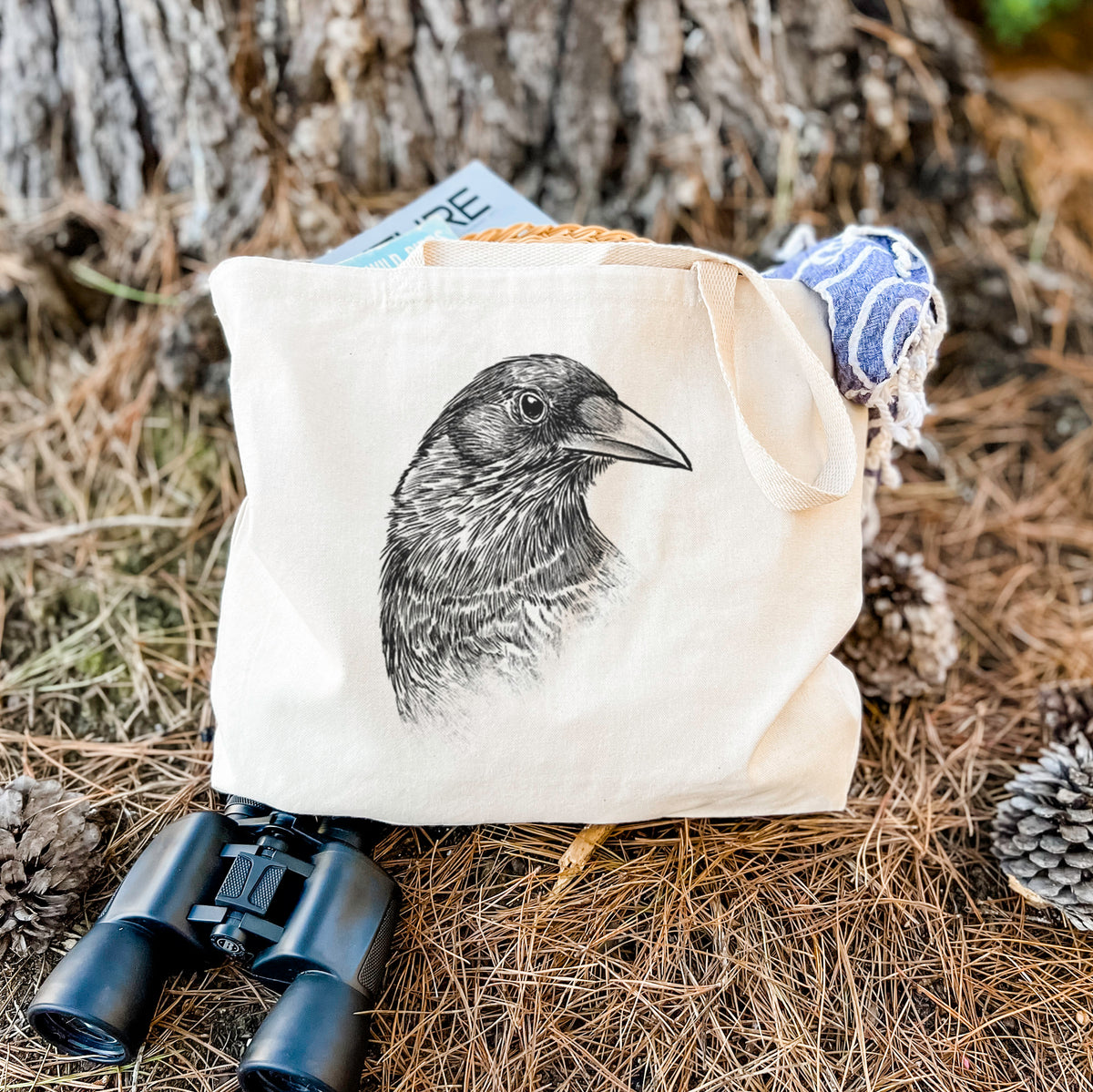 Canvas tote bag printed with a detailed black ink portrait of a crow's head in profile, resting on dry grass beside a pair of binoculars.