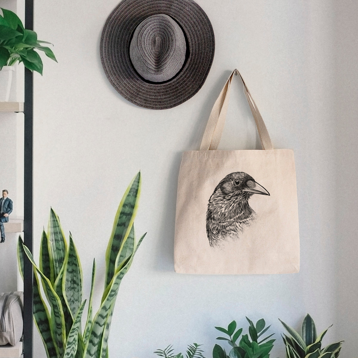 Canvas tote bag printed with a detailed black ink portrait of a crow's head in profile, hanging on a wall hook below a hat, next to potted plants.