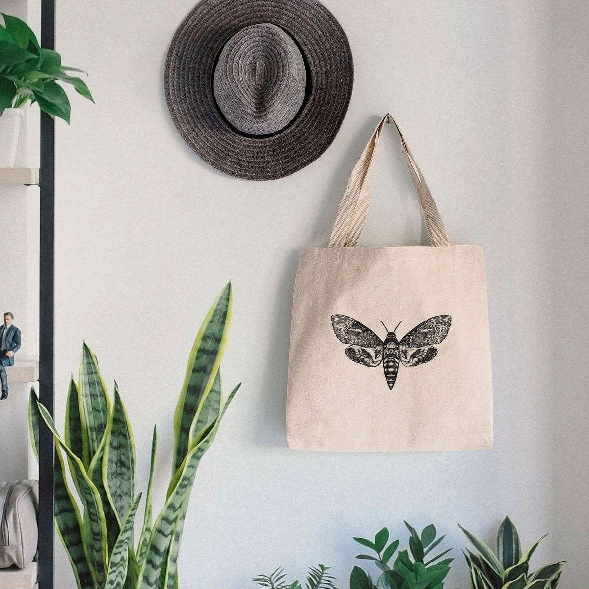 Canvas tote bag printed with a detailed black ink illustration of a death's-head hawkmoth with its wings spread, hanging on a wall hook below a hat, next to potted plants.