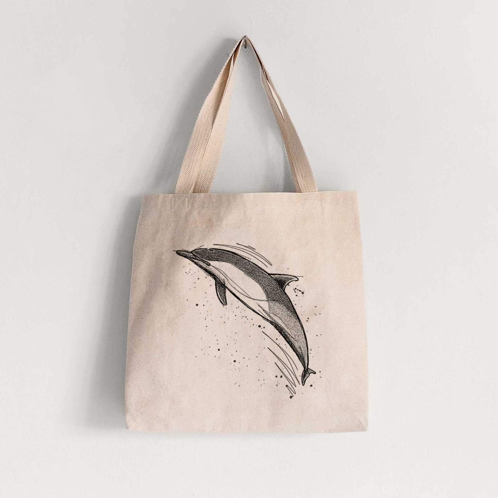 Natural canvas tote bag printed with a black ink illustration of a common dolphin leaping, with ink splatters.