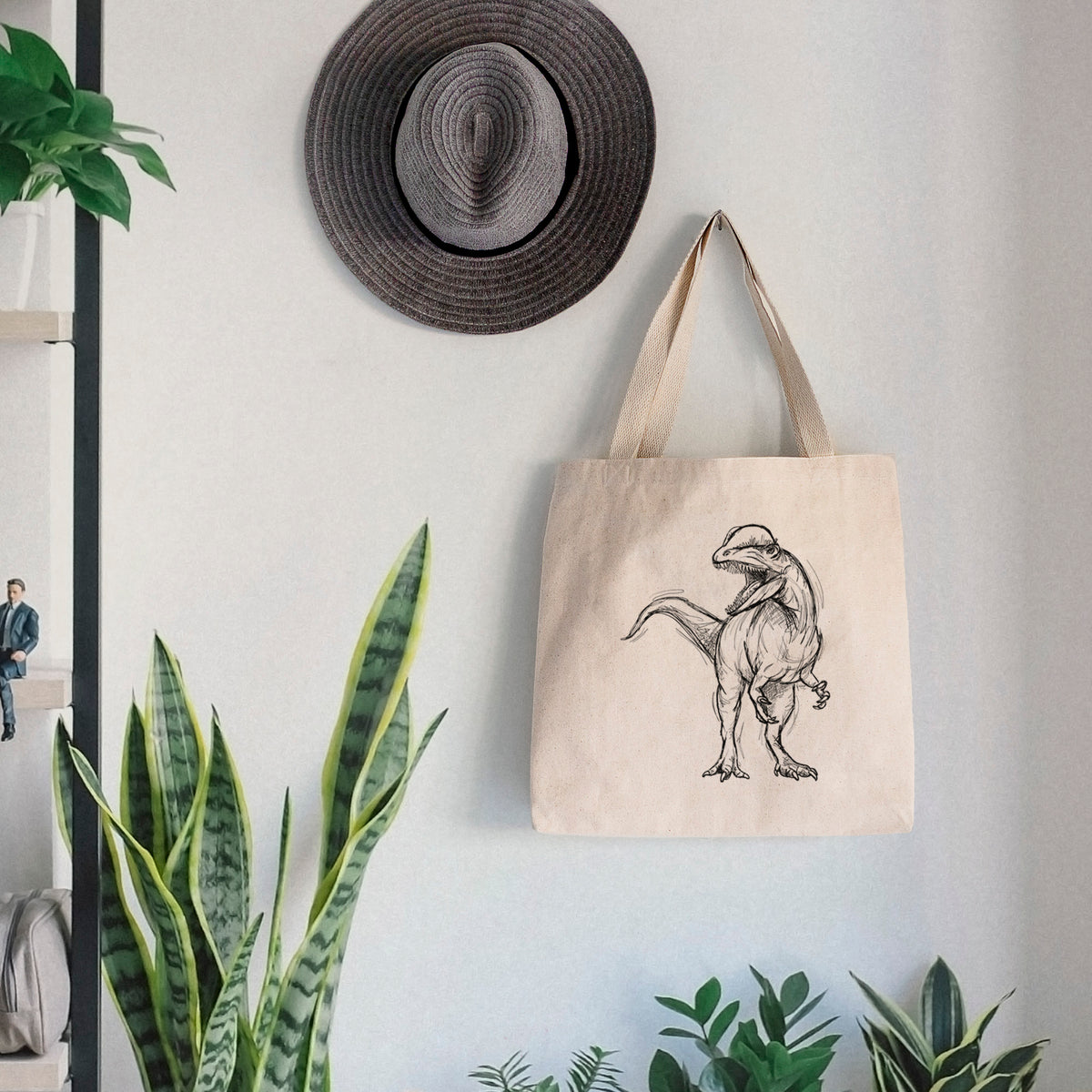 Canvas tote bag printed with a black ink illustration of a Dilophosaurus with its neck frill, walking, hanging on a wall hook below a hat, next to potted plants.
