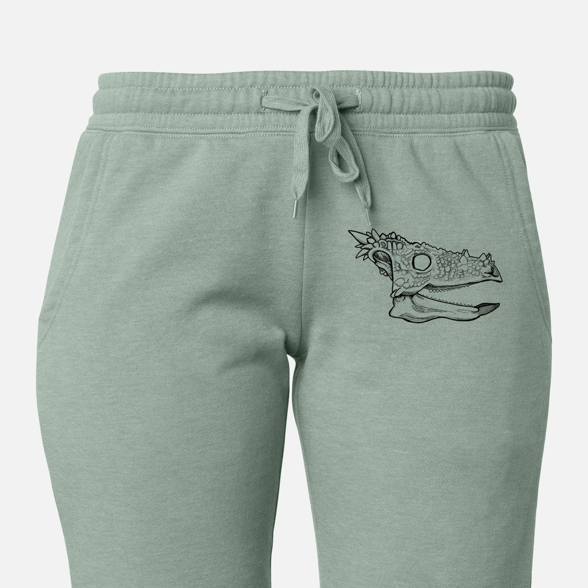 Front of sage green Cali Wave sweatpants, printed with a black ink illustration of a spiky Dracorex dinosaur skull on the leg.