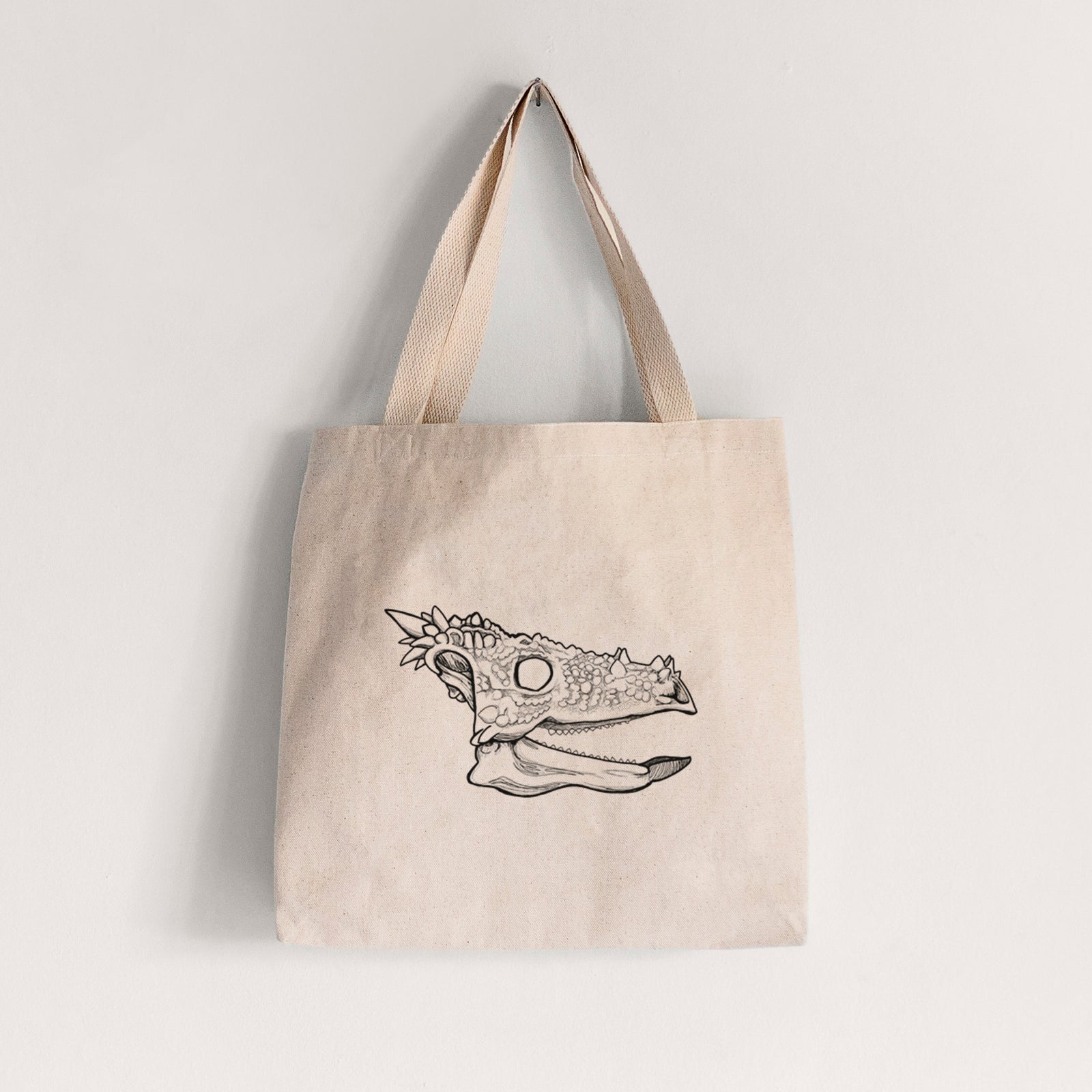 Natural canvas tote bag printed with a black ink illustration of a spiky Dracorex dinosaur skull.