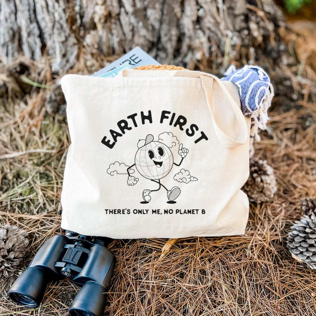 Canvas tote bag printed with a cheerful cartoon Earth running, with the words Earth First and There's Only Me, No Planet B, resting on dry grass beside a pair of binoculars.