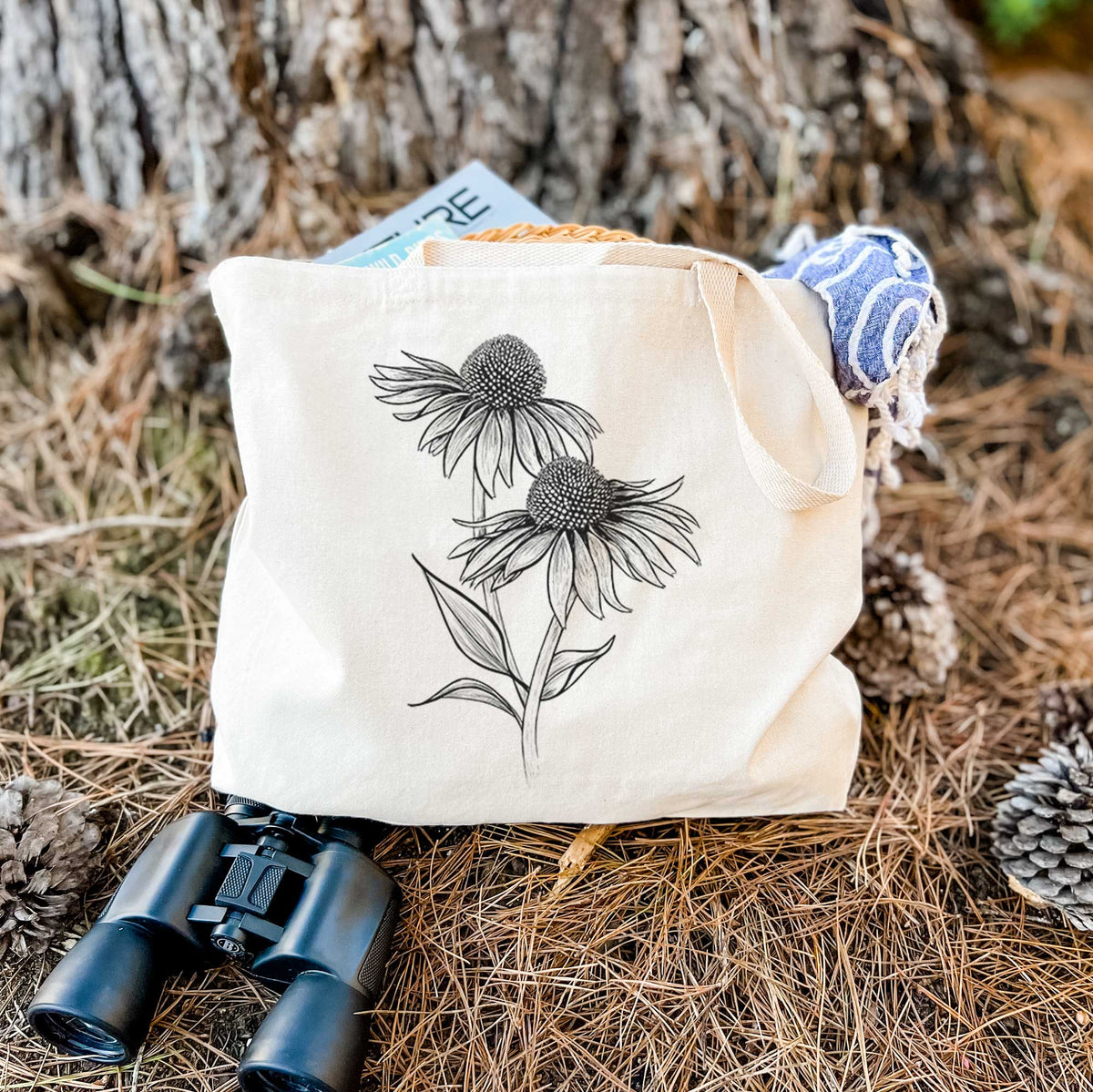 Canvas tote bag printed with a black ink line drawing of two coneflowers on tall stems, resting on dry grass beside a pair of binoculars.