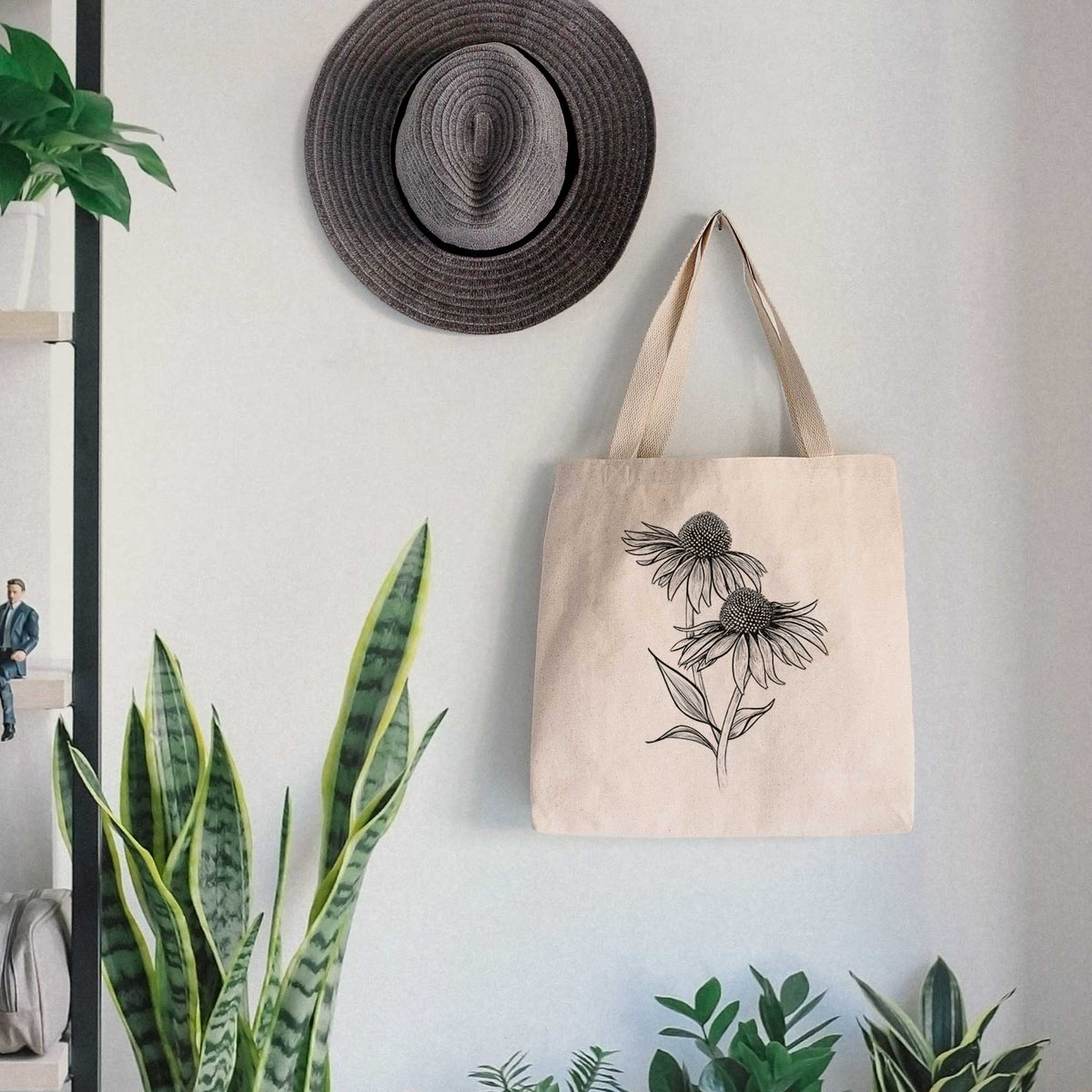 Canvas tote bag printed with a black ink line drawing of two coneflowers on tall stems, hanging on a wall hook below a hat, next to potted plants.
