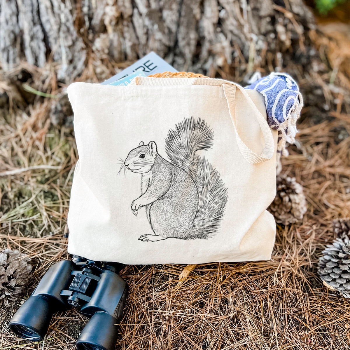 Canvas tote bag printed with a detailed black ink illustration of a fox squirrel sitting with a bushy tail, resting on dry grass beside a pair of binoculars.
