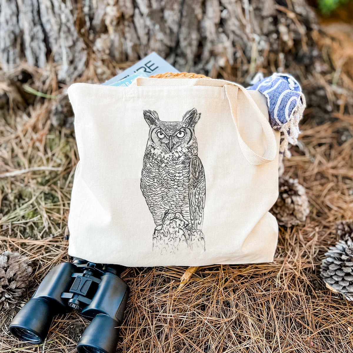 Canvas tote bag printed with a detailed black ink illustration of a great horned owl standing upright with its ear tufts raised, resting on dry grass beside a pair of binoculars.