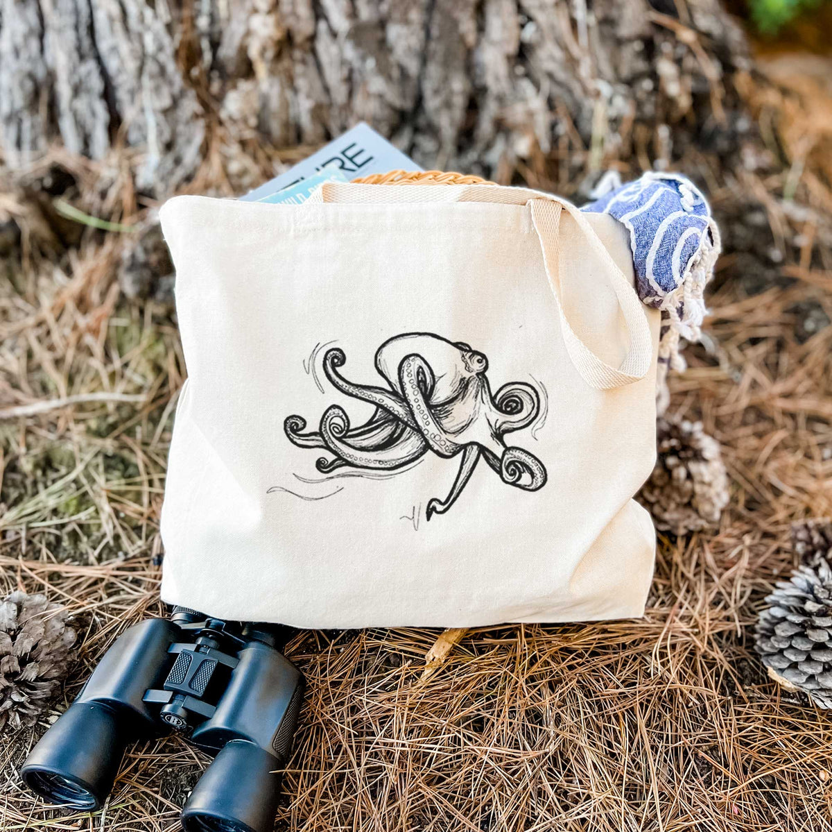 Canvas tote bag printed with a black ink illustration of a giant Pacific octopus with curling tentacles, resting on dry grass beside a pair of binoculars.