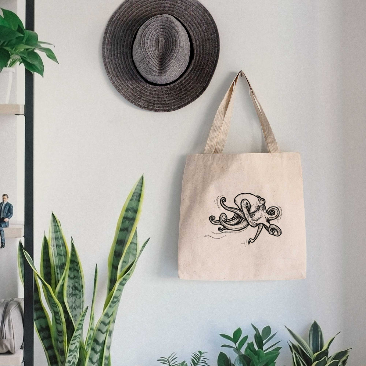 Canvas tote bag printed with a black ink illustration of a giant Pacific octopus with curling tentacles, hanging on a wall hook below a hat, next to potted plants.
