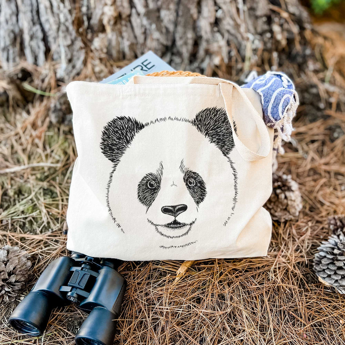 Canvas tote bag printed with a black ink portrait of a smiling giant panda's face, resting on dry grass beside a pair of binoculars.