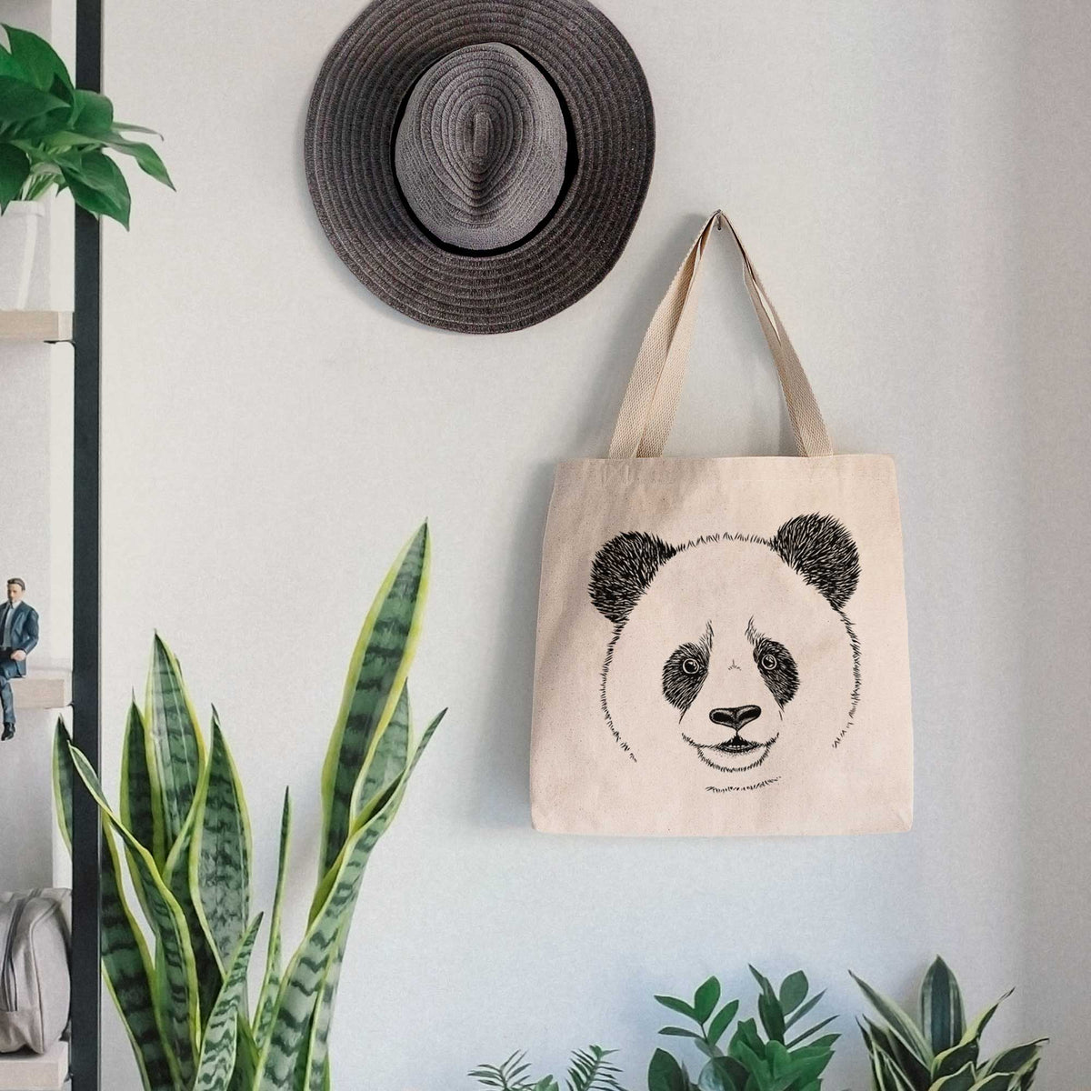 Canvas tote bag printed with a black ink portrait of a smiling giant panda's face, hanging on a wall hook below a hat, next to potted plants.
