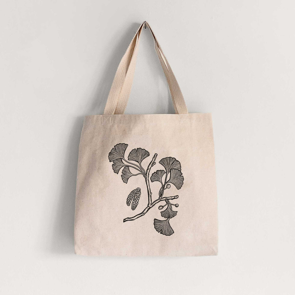 Natural canvas tote bag printed with a black ink illustration of a ginkgo branch with fan-shaped leaves.