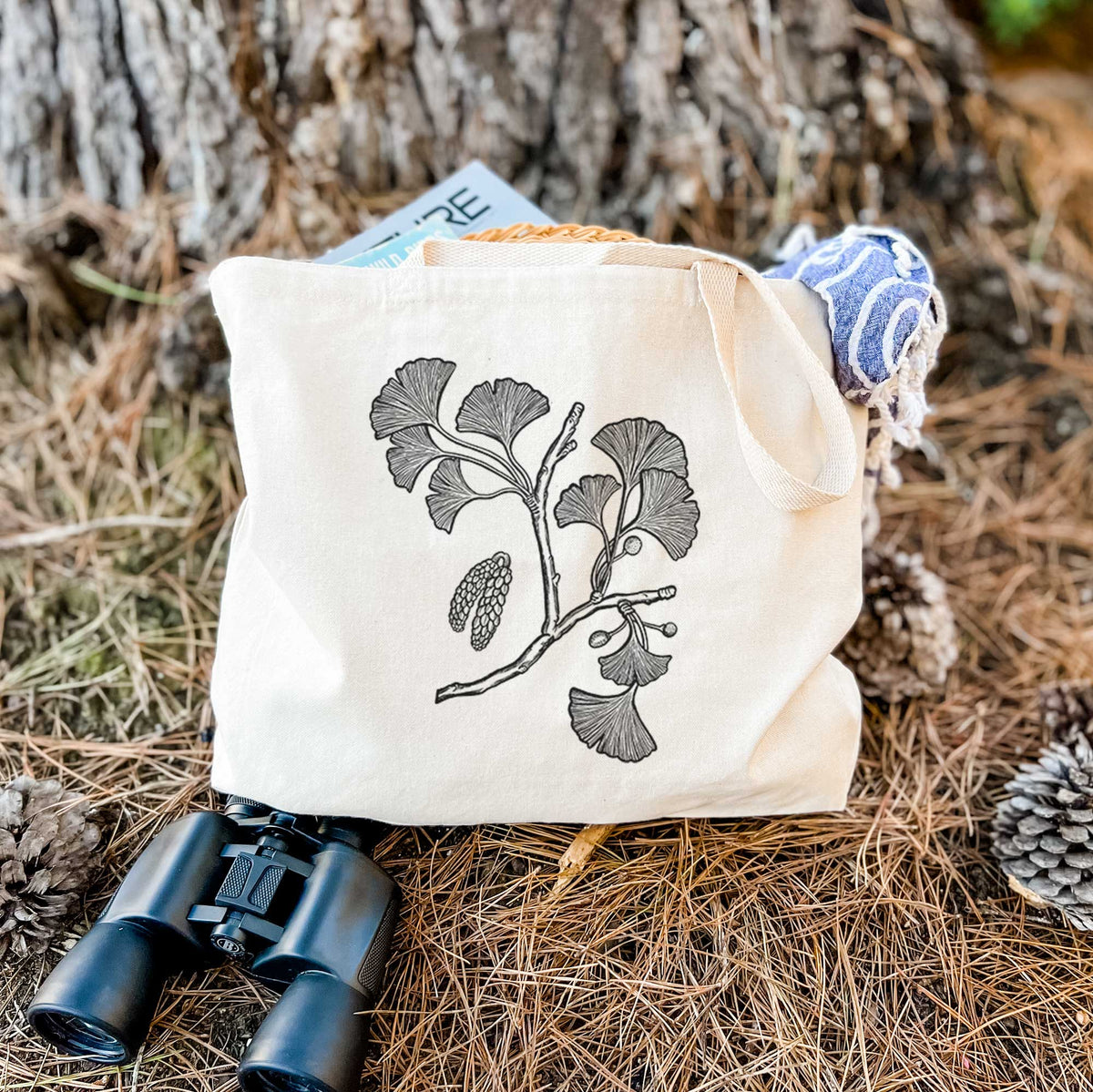 Canvas tote bag printed with a black ink illustration of a ginkgo branch with fan-shaped leaves, resting on dry grass beside a pair of binoculars.