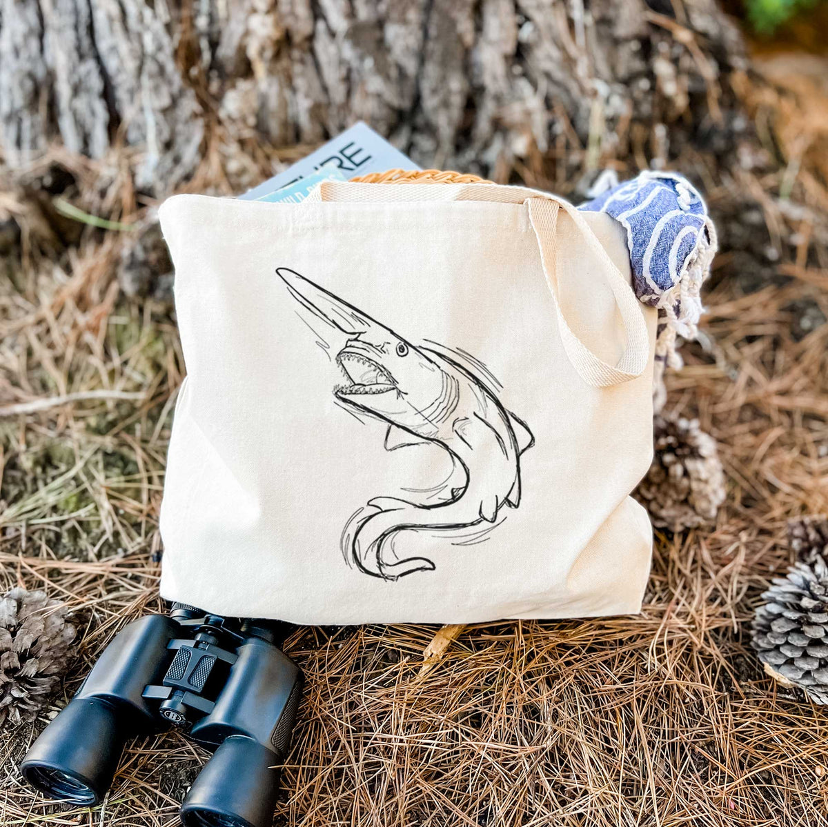 Canvas tote bag printed with a black ink illustration of a goblin shark with its long snout, swimming in a curve, resting on dry grass beside a pair of binoculars.