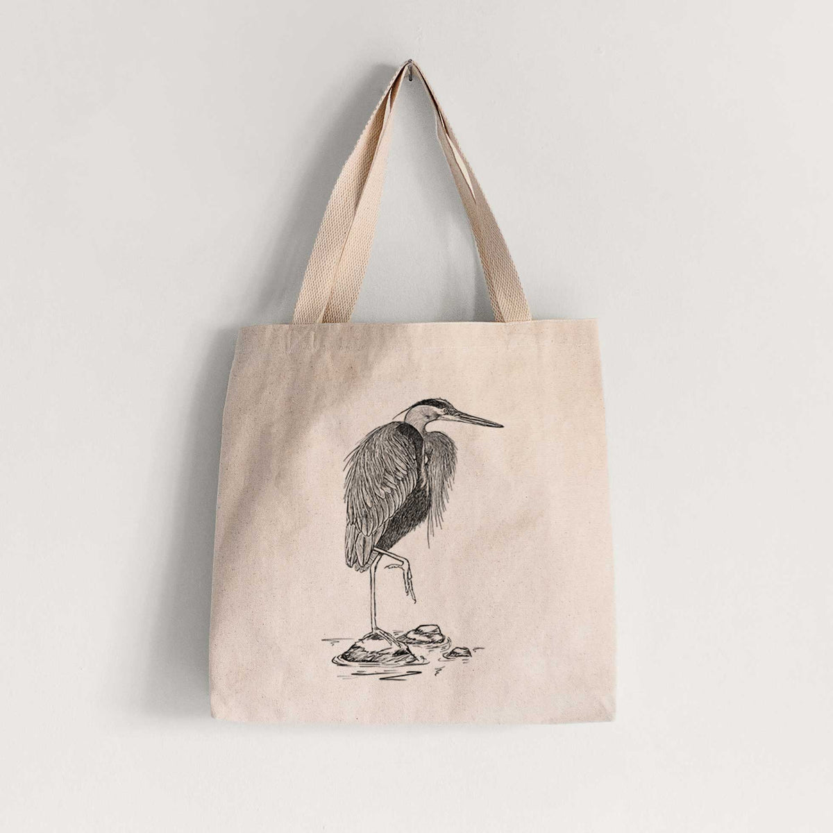 Natural canvas tote bag printed with a detailed black ink illustration of a great blue heron standing on rocks.