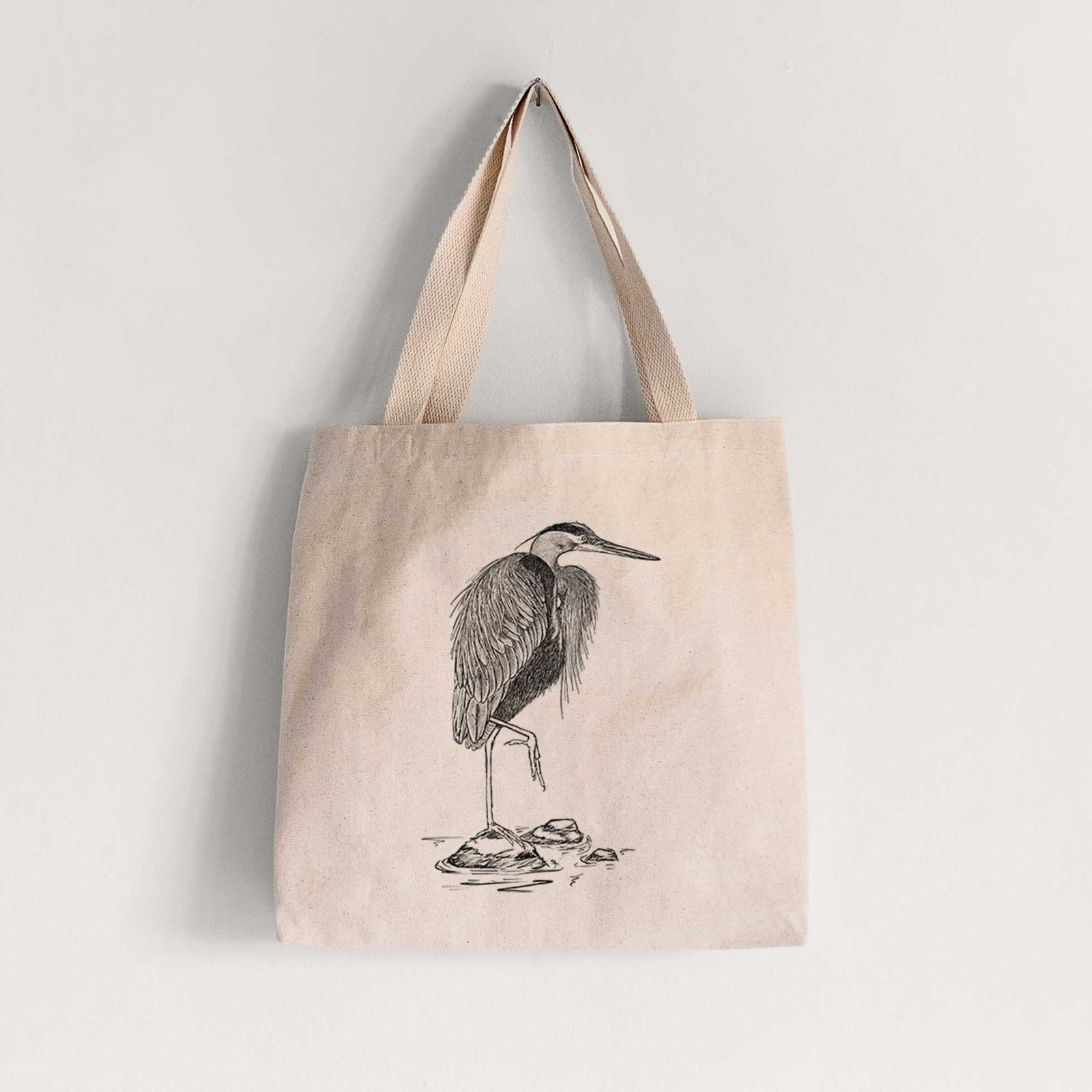 Natural canvas tote bag printed with a detailed black ink illustration of a great blue heron standing on rocks.