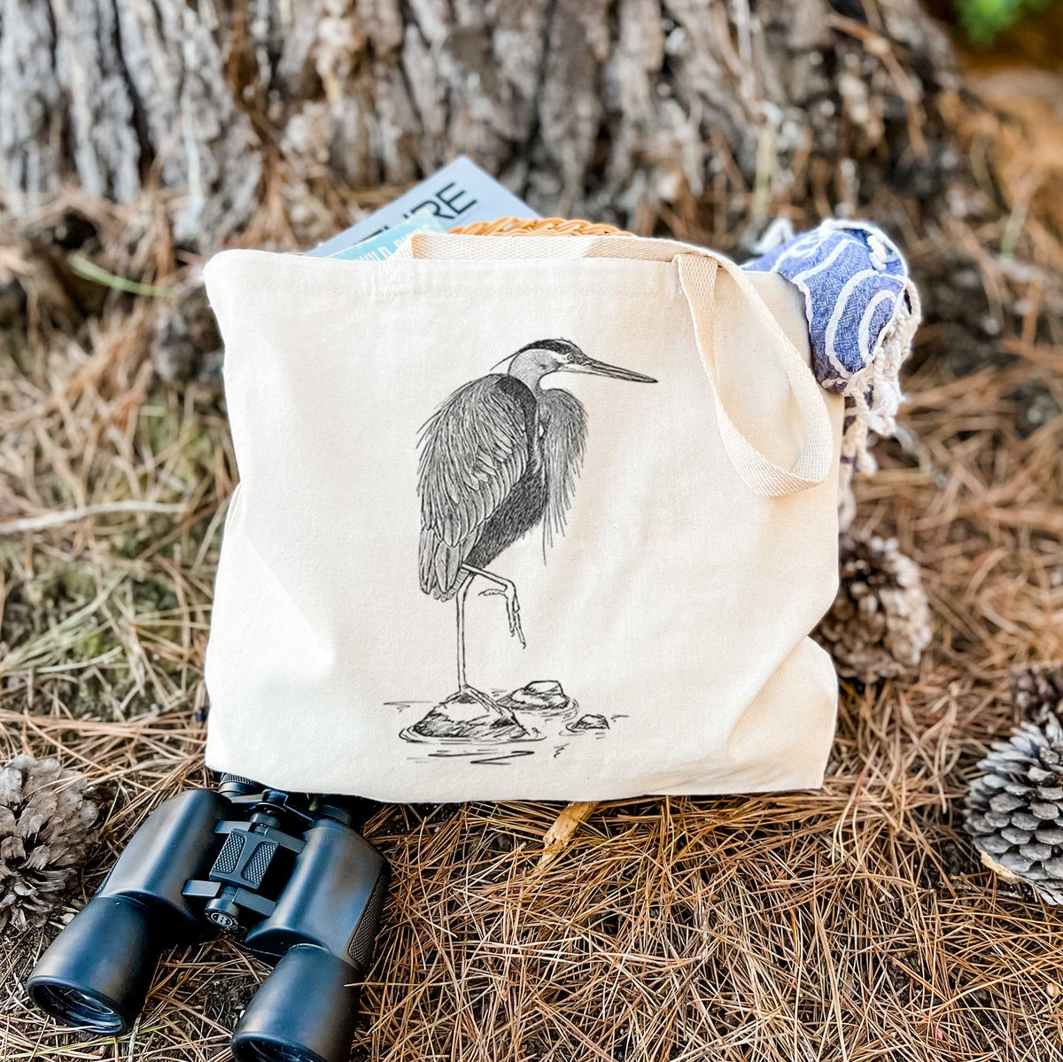 Canvas tote bag printed with a detailed black ink illustration of a great blue heron standing on rocks, resting on dry grass beside a pair of binoculars.