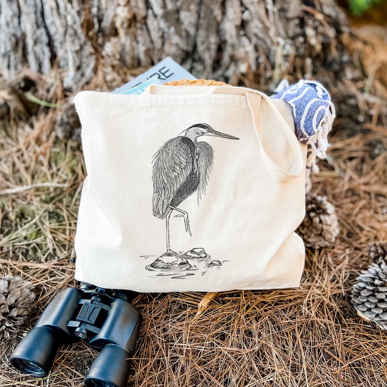 Natural canvas tote bag printed with a detailed black ink illustration of a great blue heron standing on rocks.