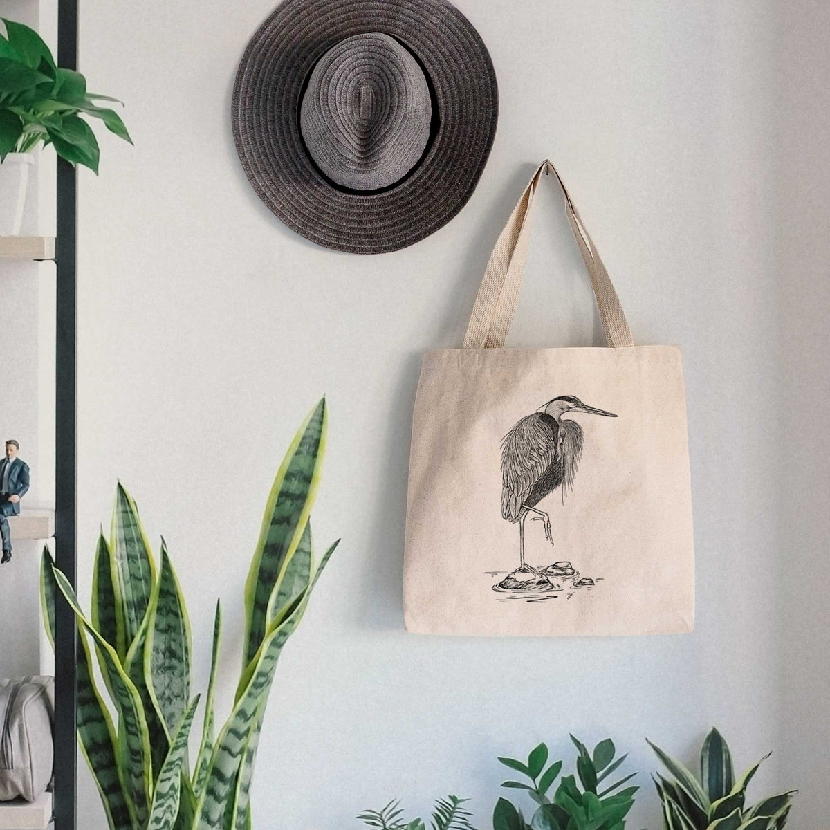 Canvas tote bag printed with a detailed black ink illustration of a great blue heron standing on rocks, hanging on a wall hook below a hat, next to potted plants.